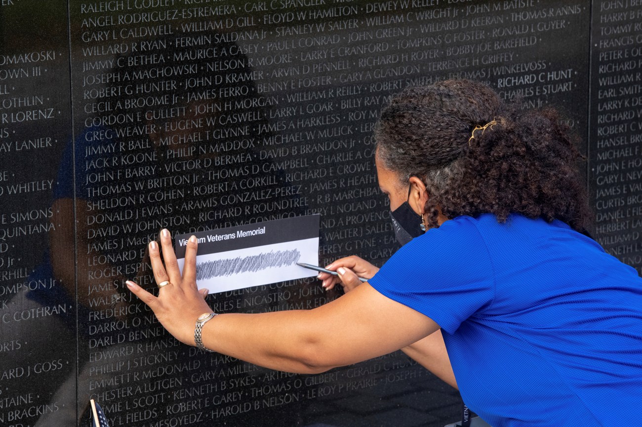 A woman in bullet uses a pencil to rub a name from the Vietnam Veterans Memorial Wall onto paper.