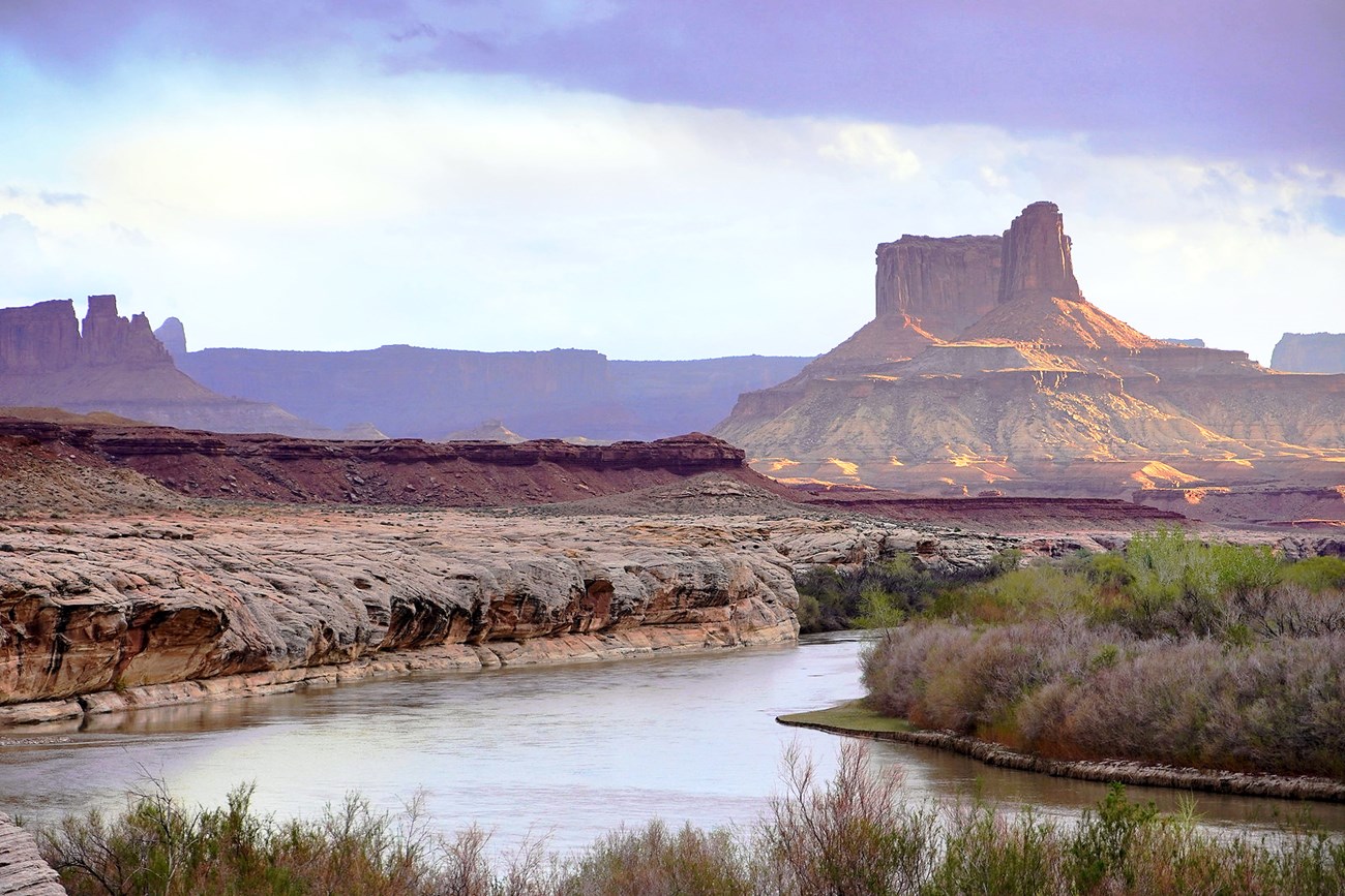 Green River Calm section of river, lined with sheer rock on one side and dense shrubs on the other. Behind it, low, golden-hour sunlight strikes a stunning rock formation from one side, while more distant shaded plateaus appear purple.
