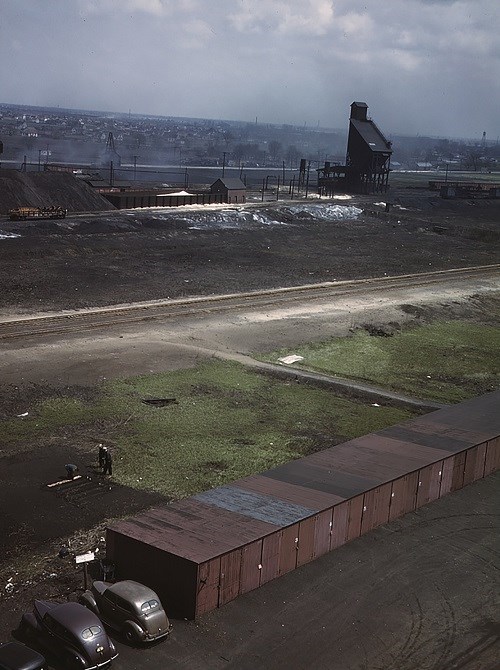 Color photo overlooking a rail yard. In the lower portion are two cars parked next to a long row of sheds. In an adjacent grassy area, a garden bed has been tilled and partly planted. In the distance is more rail yard and then homes under an overcast sky.