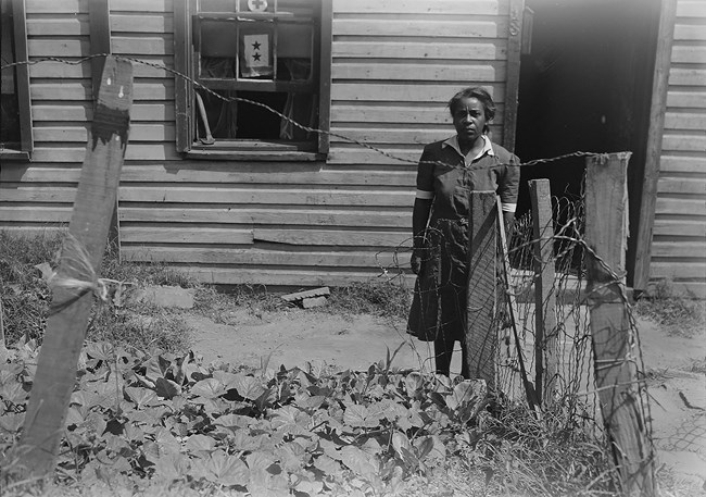 Black and white photo of a Black woman in a dress standing in front of a clapboard row house. The door is open. She is standing by a garden in her front yard. The window is open.