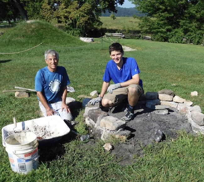 Two peoploe crouch down near a partially constructed fire ring.