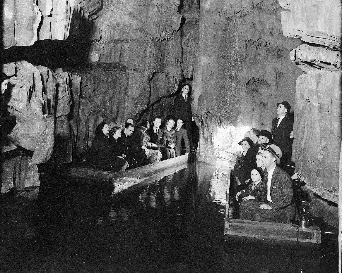 Two small wooden boats filled with people wearing suits and dresses sit on a small body of water surrounded by jagged cave walls.