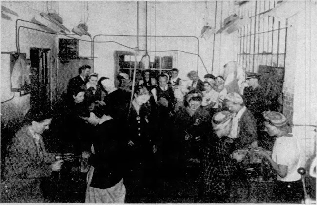 Pensacola News Journal. 1944. Black and white grainy photograph of groups of women standing over devices on tables. Three men stand in the crowd.