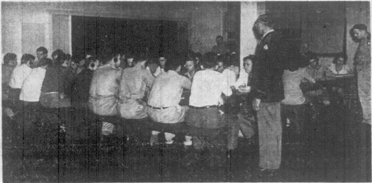Pensacola New Journal. 1944. Black and white photo of men seated at a long table with their backs to the camera wearing headphones. A man in uniform stands at one end of the table