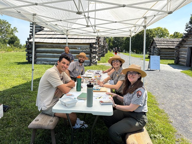 smiling park rangers sit at a table with a smiling intern near log huts