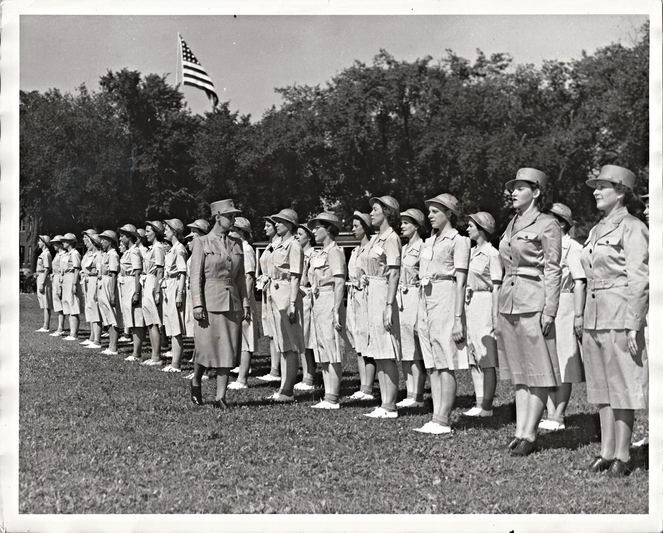 Women in uniforms (collared, light-color dresses) and round, brimmed hats stand at attention side by side for inspection on a grassy lawn. Tops of trees and an American flag are visible in the background