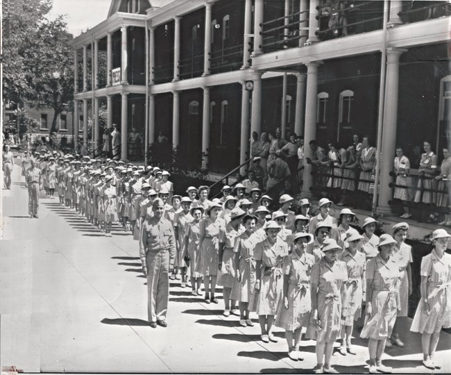 Women in uniforms (light-color collared dresses) and round brimmed hats stand in three lines to form ranks in front of barrack buildings. Men and women crowd the porch of the buildings behind a railing