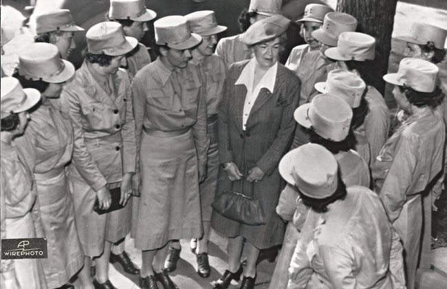 Women wearing cylindrical military caps and collared dresses with skirts below the knee stand side by side in a U shape to meet an older woman. The woman wears a dark, pin-striped dress with a white collared shirt and holds a small purpose.
