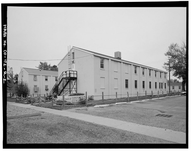 Perspective view of long, two-story barrack building with smooth, light-colored walls and a pitched metal roof. Metal stairs descend from a door on the second floor to the ground on the short side of the building