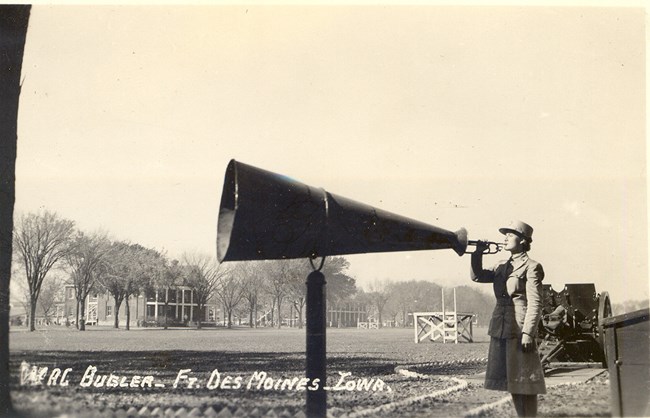 Uniformed woman plays bugle into giant, free-standing megaphone. She is standing in a large field. Two houses, trees, a wooden platform, and a tractor are visible in the background