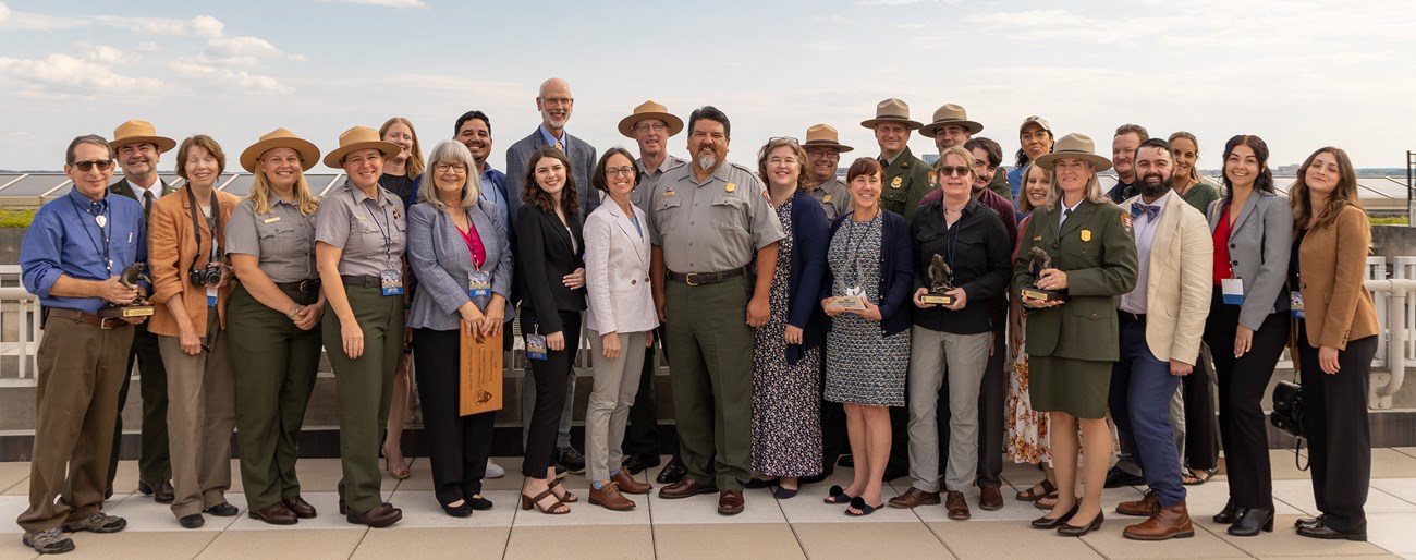 A couple dozen people wearing ranger uniforms or buisness attire pose for a photo on a rooftop.