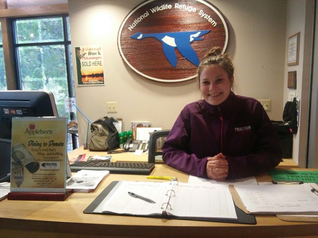 A brunette woman smiles from behind the front desk of a Visitor Center. A wooden sign behind her identifies it as a part of the National Wildlife Refuge System and she is wearing a purple jacket reading “Friends of Sullys Hill.”