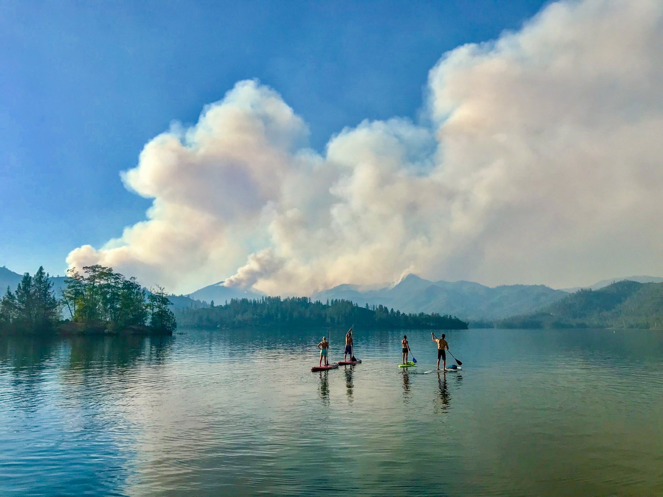 Four people paddleboarding on a calm lake, looking towards the billowing smoke of the Carr Fire raging in the distance.