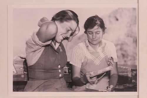 Elzada Clover and Lois Jotter sit next to each other looking at a book. Elzada Clover and Lois Jotter sit next to each other looking at a book.