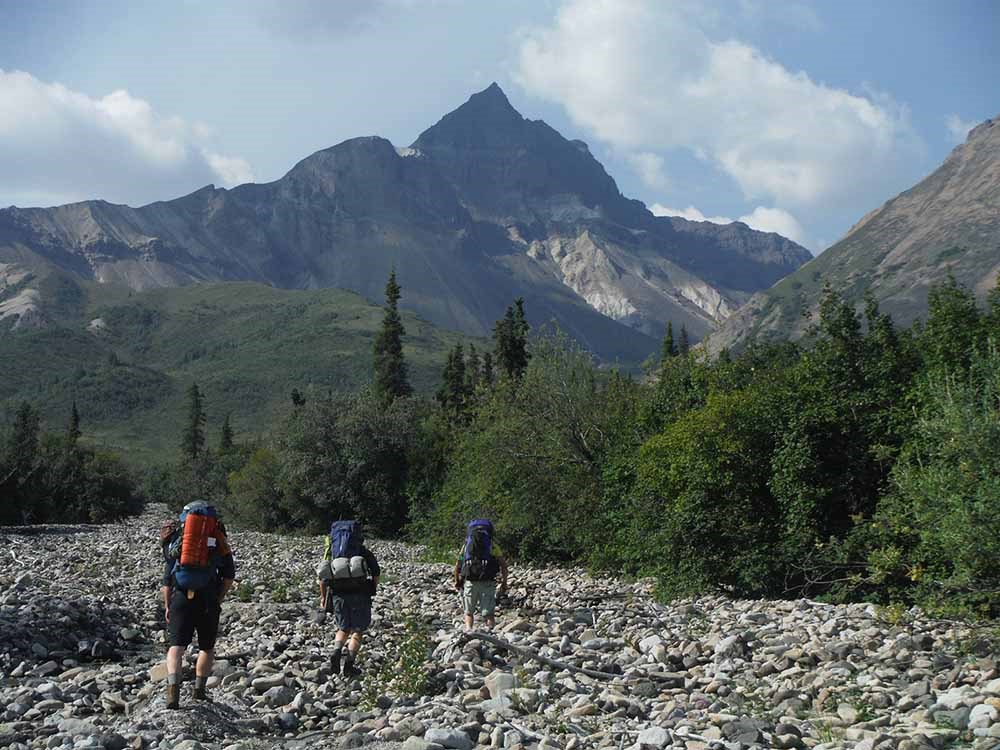 Three men with backpacks hike a boulder-strewn dry creek bed into the mountains.