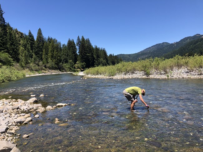 A man bends over to collect a water sample in a shallow mountain river