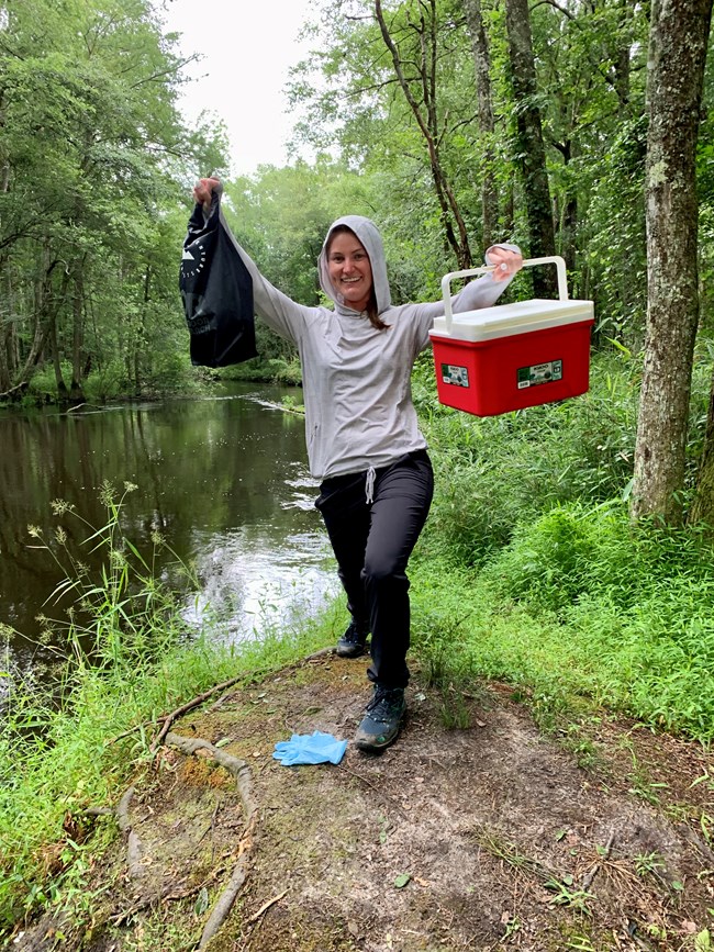A woman stands on a river band holding up a red cooler and black drybag used for collecting water samples