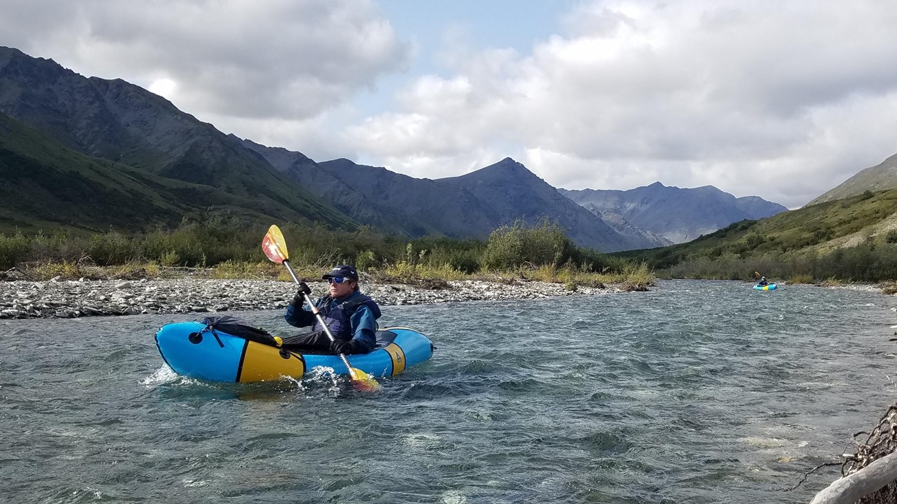 A person in an inflatable kayak paddles down a river with mountains in the background