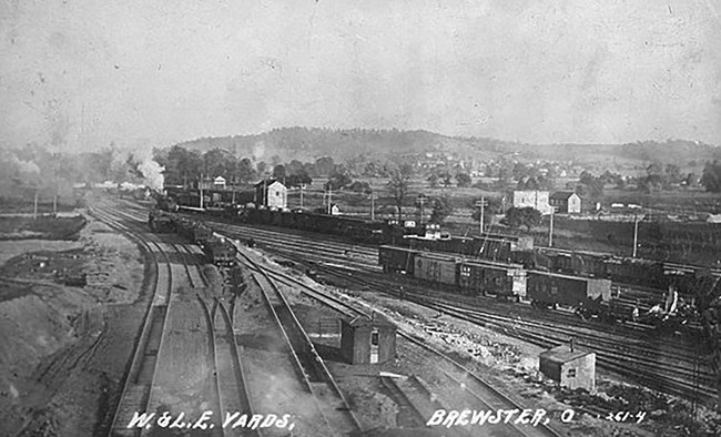 Black and white photo postcard overlooking a busy rail yard. There are several tracks, switches, and small buildings. Lines of rail cars sit on the tracks. In the distance are low mountains.