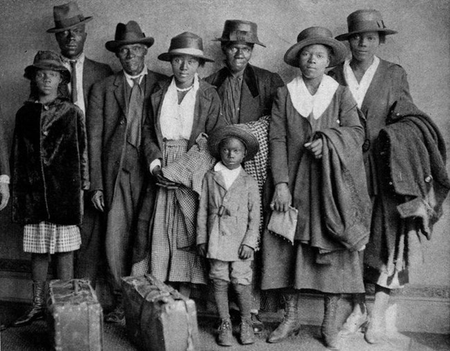 Black and white photo of a Black family (two men, five women, and a young boy) dressed in suits and dresses, all wearing hats. In front of them are two suitcases.