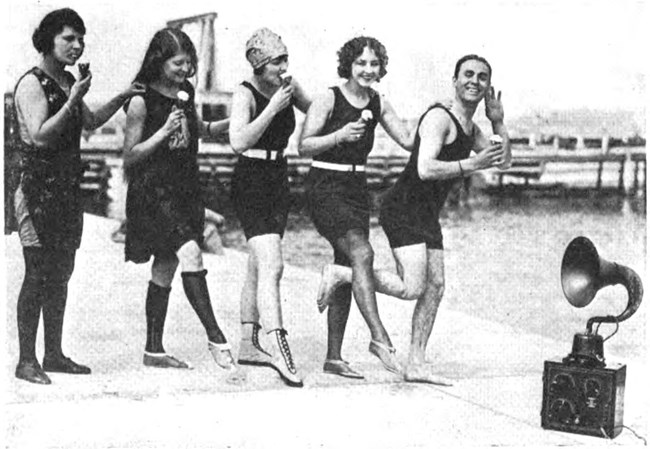 Black and white photo pf four white women and a white man wearing 1920s bathing suits and eating ice cream cones. A box with knobs and a trumpet-shaped amplifier (the radio) sits on the beach in front of them.