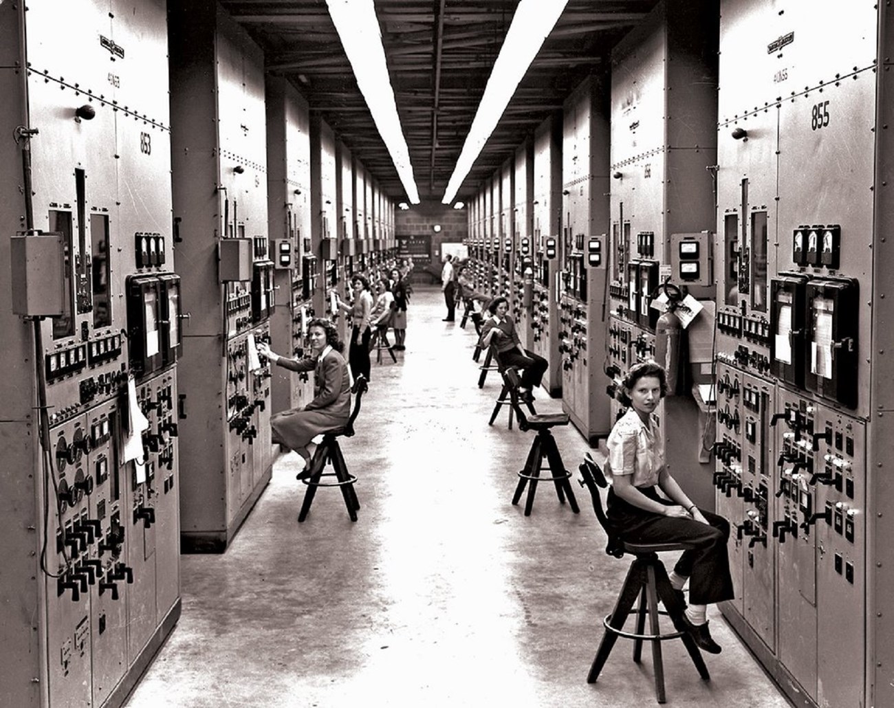 lt= Women sitting on stools in front of large mainframe computer s that reach to the ceilings. There's a long aisle between the computer banks.