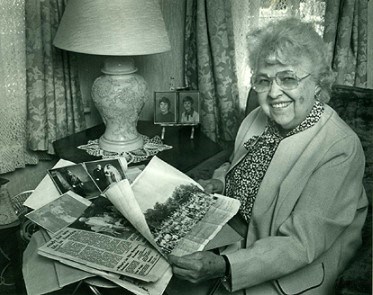 Black and white photo of older woman with glasses looking through old newspapers and photos.