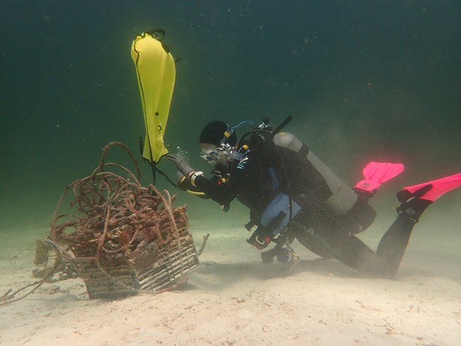 A scuba diver on the right attached a bright yellow lift bag to a large rectangular lobster trap laden with thick fishing line. The lift bag will be filled with air to bring the marine debris to the surface.