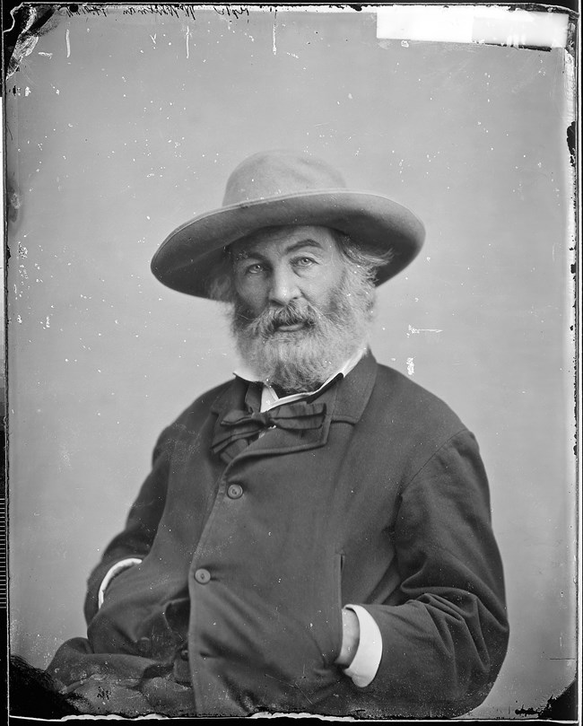 Seated photograph of Walt Whitman in straw hat with long beard.