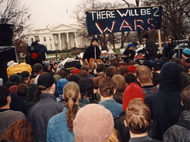 Fugazi performing in front of the White House on the eve of the Persian Gulf War. 1991. a crowd gathers in front of a band performing in front of the white house, under a banner that says "there will be 2 wars"