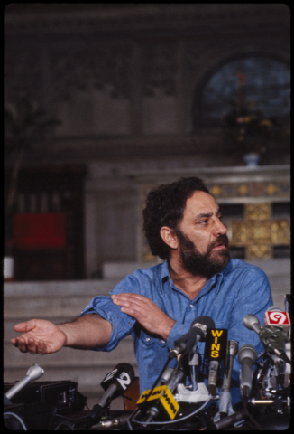 A bearded man seated in front of a bunch of microphones