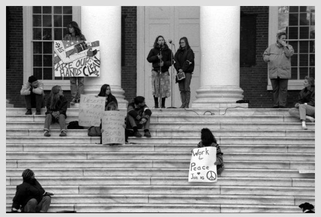 Two women stand and speak into a microphone at the top of some steps, while several more sit holding signs in front of them. Signs say "Work for peace, join us", "you cant see a thousand points of light from a bomb shelter", and "our hands are clean"