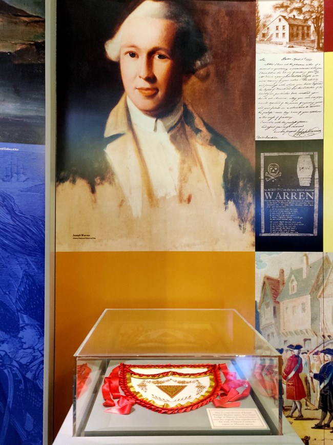 Exhibit display with a portrait of Joseph Warren on wall and a Masonic Apron laying flat in a glass display case.
