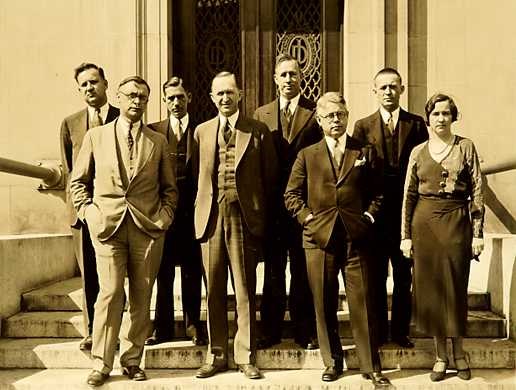 Seven men wearing suits, ties and vests and one woman wearing a ankle length skirt stand on stairs in front of an elaborately carved door that stands partially open.