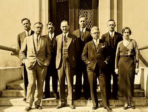 Seven men wearing suits, ties and vests and one woman wearing a ankle length skirt stand on stairs in front of an elaborately carved door that stands partially open. Seven men wearing suits, ties and vests and one woman wearing a ankle length skirt stand on stairs in front of an elaborately carved door that stands partially open.