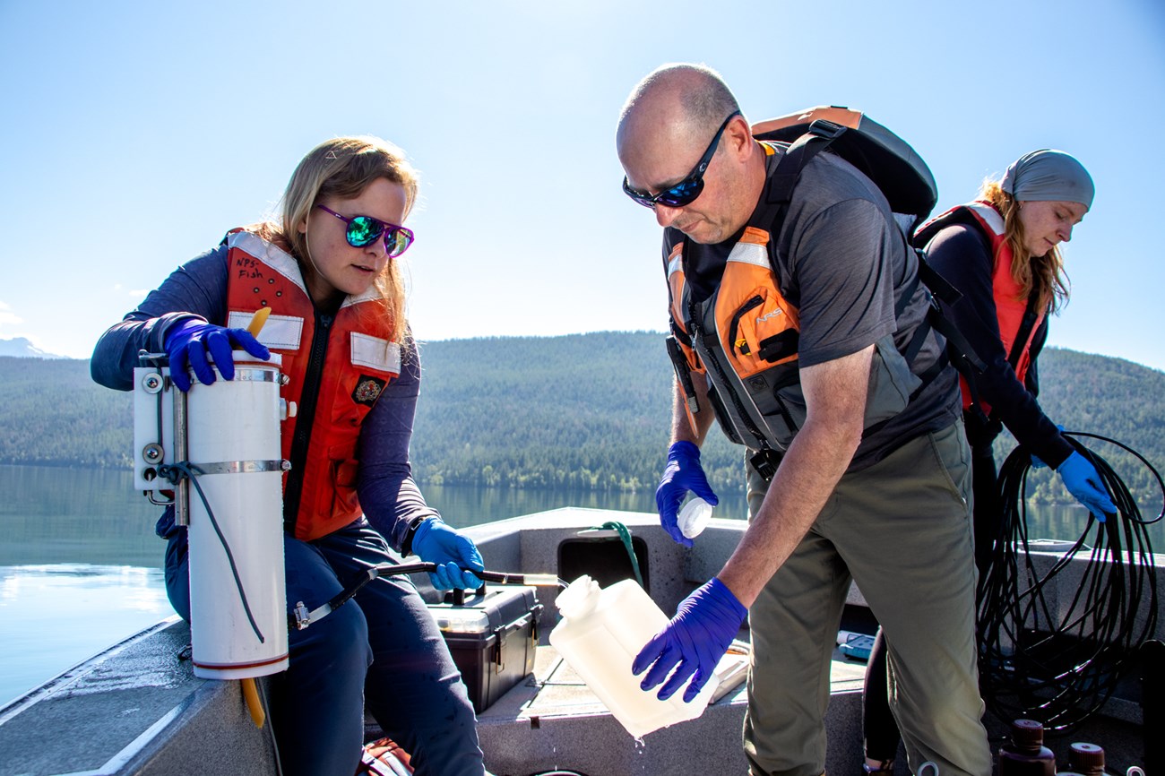 Three people on a boat hold containers and hoses used to collect water samples and water quality sample measurements.