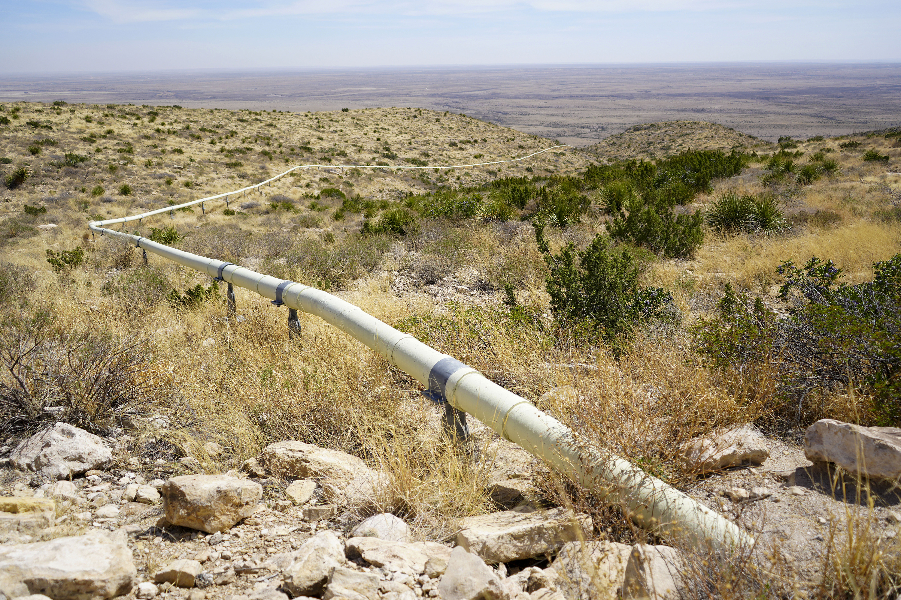 Photo of mountain slope with dry grass, shrubby evergreens, rocks, and a white pipe snaking up it.