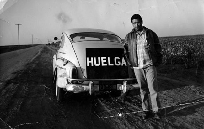 César Chávez standing with car with the word "Huelga" (strike) on the back.