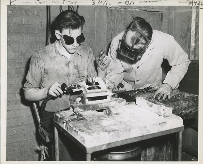 Black and white photo of a young white man wearing welding goggles sitting at a work bench to weld. Another white man, wearing a welding mask, bends over the table besides him to inspect his work.