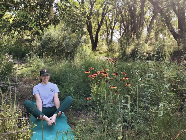 A smiling Park Ranger demonstrates flower pose while sitting on a yoga mat in a pollinator garden with blooming flowers. A smiling Park Ranger demonstrates flower pose while sitting on a yoga mat in a pollinator garden with blooming flowers.