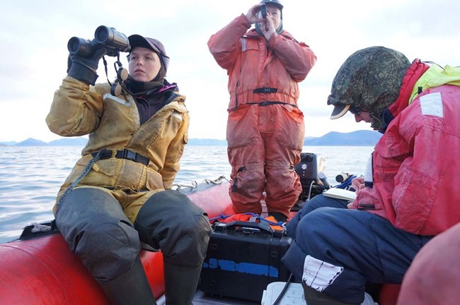 Three people in an inflatable boat. One records data, one looks through binoculars, and the other looks through a range finder.