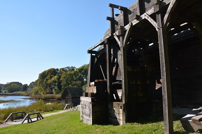 wooden structure with large wheel on a hill overlooking a river under a blue sky.