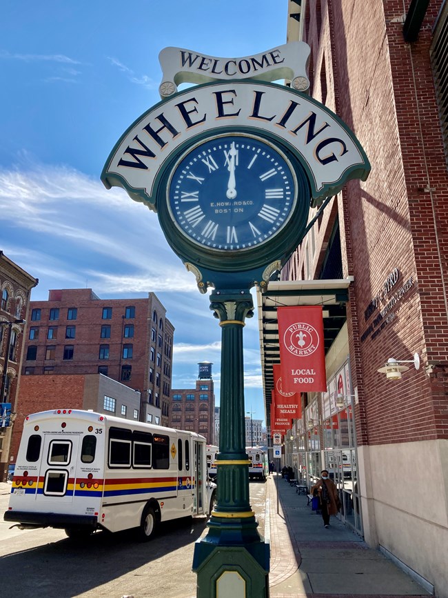 Large, decorative old-fashioned analog clock on tall post. Engraved text on ribbon motif above clock face reads 'Welcome - Wheeling'. Brick buildings, city street, and small public bus in background.