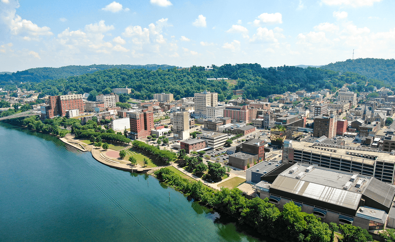 Wheeling, WV 'Bird's eye' perspective of river in foreground with tree-lined shore, city buildings, and forested hills in background with blue. sunny sky above
