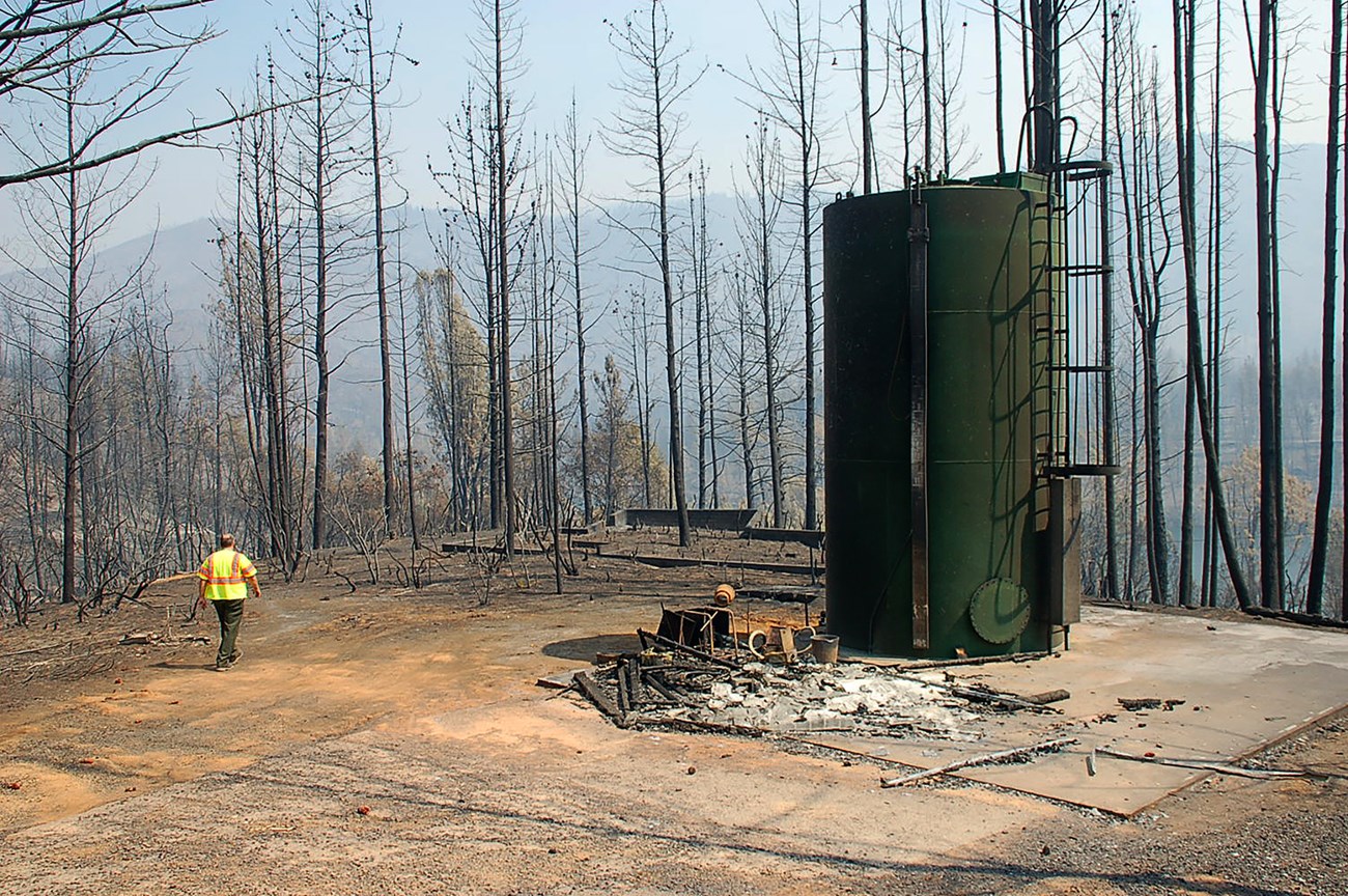 Dark green steel structure looms over a charred landscape, with a person in a bright safety vest walking among the surrounding debris..