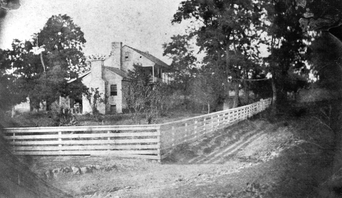 Black and white image of gray two-story frame house surrounded by trees and white fence.