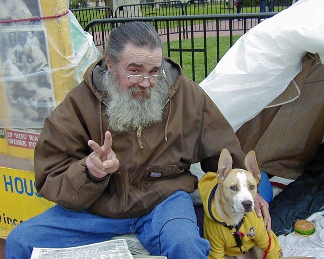 Activist William Thomas smiles and flashes a peace sign at the camera while sitting with his dog. Smiling bearded man sits with a dog while making a peace sign.