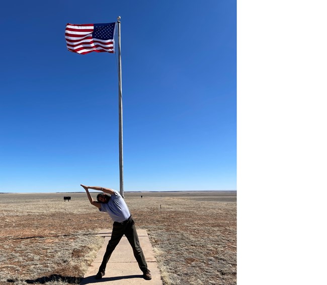 Park Ranger in yoga pose standing in front of fully extended flag on flagpole. Park Ranger is leaning to their right with both arms extend above their head and palms of each hand flat against one another.