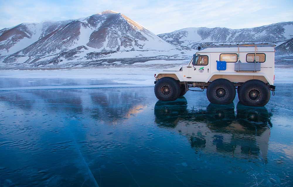 A white land rover on clear ice with snowy mountains in the background.