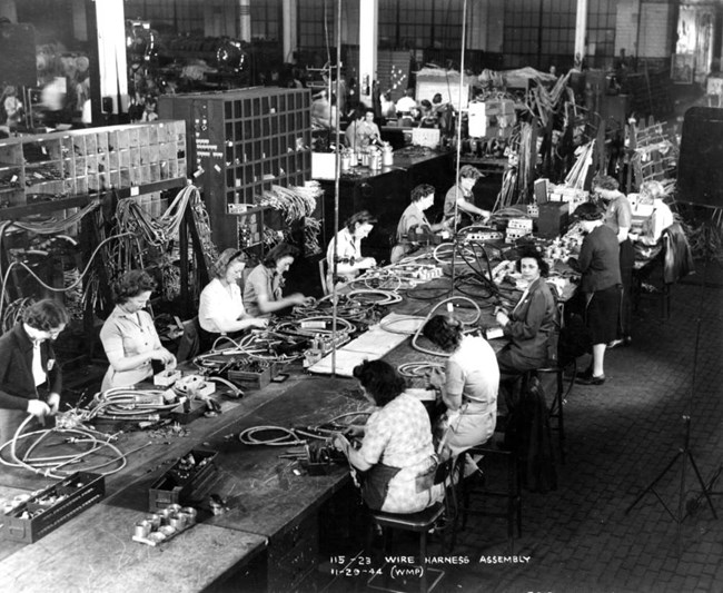 Women sit on either side of a long table in a large open room with a high ceiling. Their heads are bent to work with long pieces of thick metal cable on the table. Wires hang on shelving behind them. Other workstations are visible behind them.